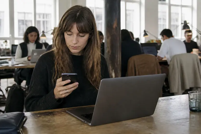 Transfert de milliers de photos Android vers PC : une femme dans un espace de coworking utilise son smartphone et son ordinateur.